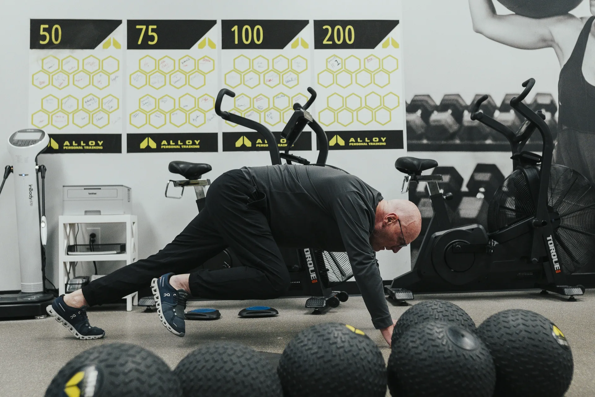 A member works through a coached floor exercise beside a row of slam balls at an Alloy Personal Training studio