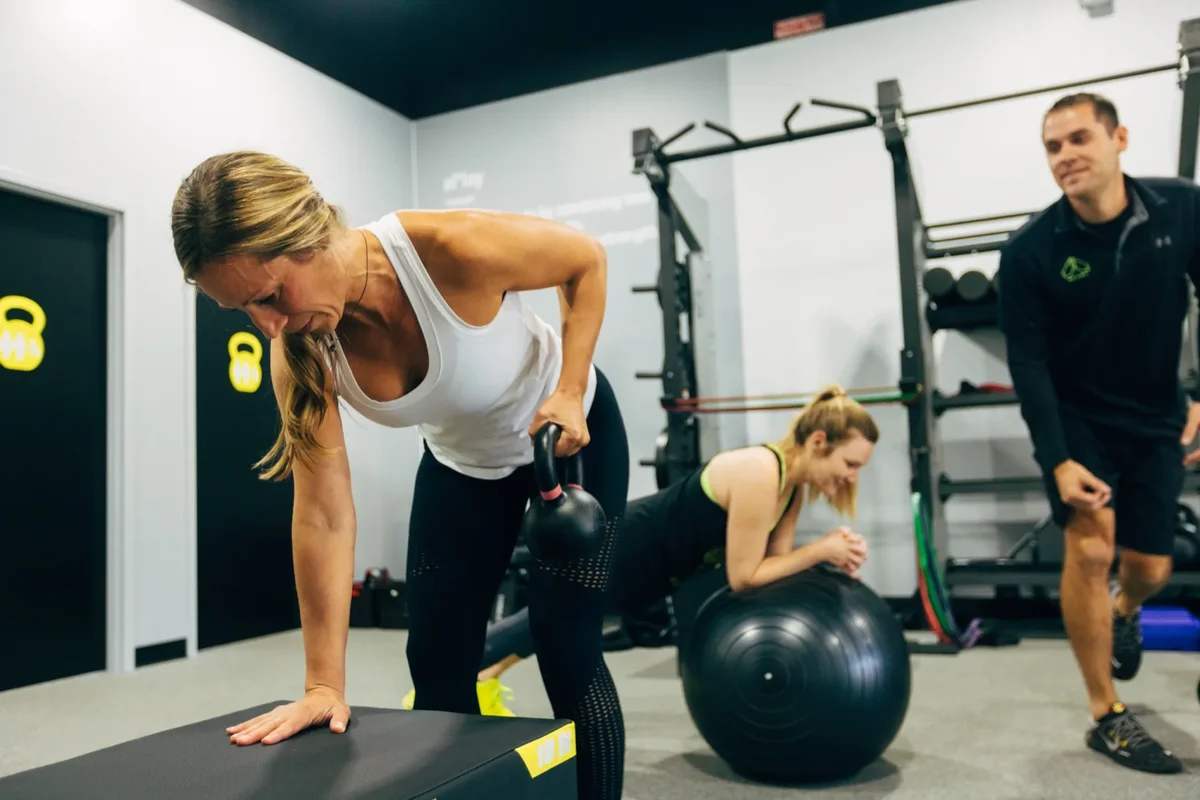 A member works through single-arm kettlebell rows while a coach leads a small group session