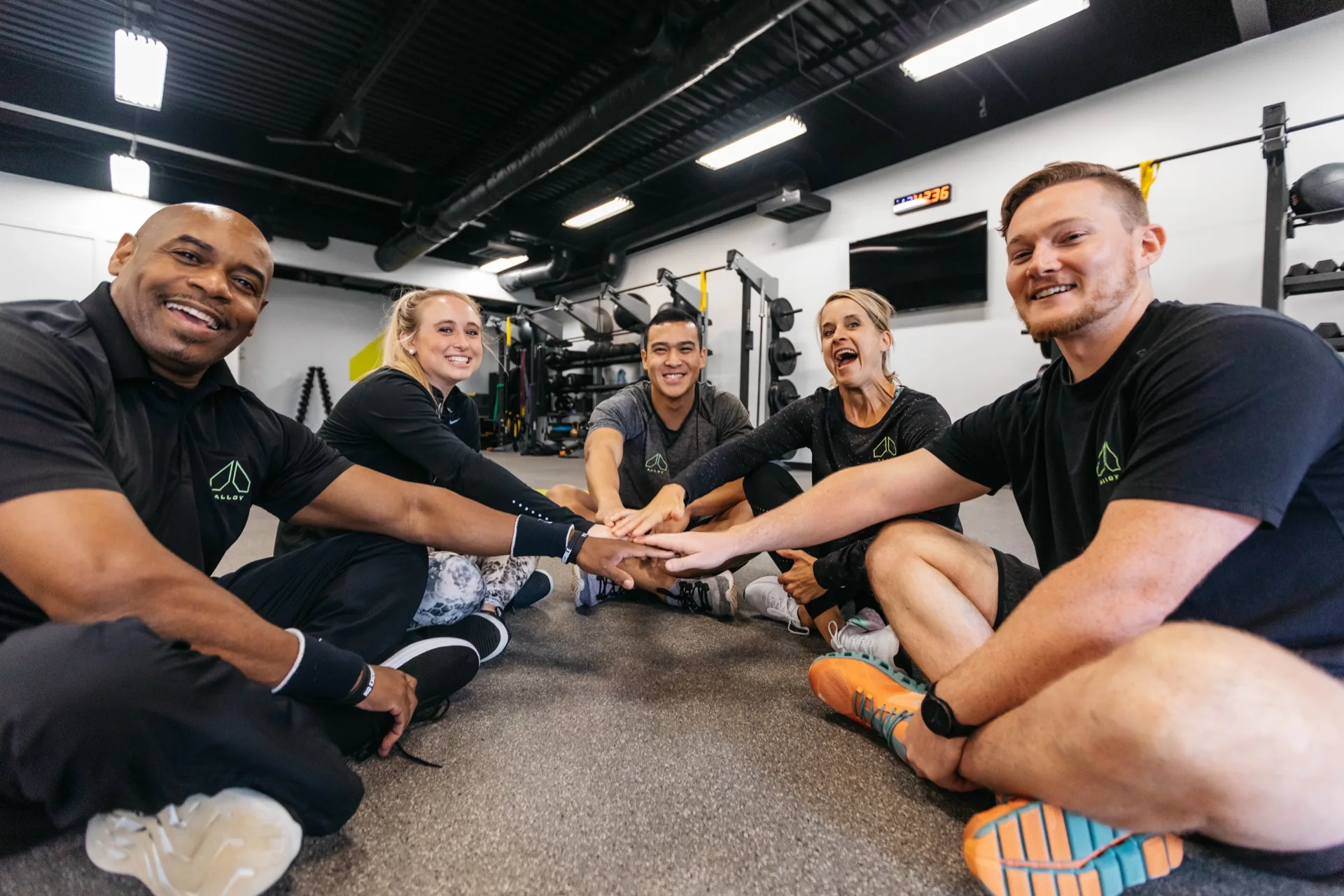 Members and coaches stack hands in a huddle on the training floor at an Alloy Personal Training studio